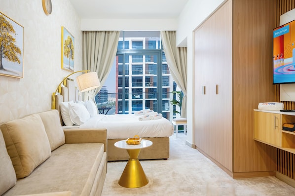 Una habitación de hotel moderna con cama, sofá, una mesa redonda dorada, televisor y un ventanal grande con cortinas.