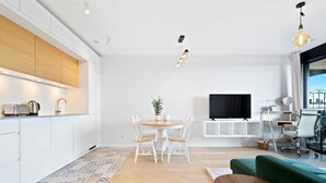 Una cuina moderna amb armaris blancs, una zona de menjador amb taula rodona i cadires, un sofà verd i un televisor penjat a la paret.