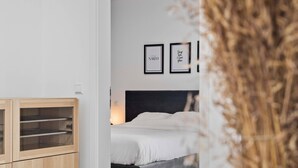 A modern room with a wooden dining table, a vase with dried plants, and a bed with a dark headboard.