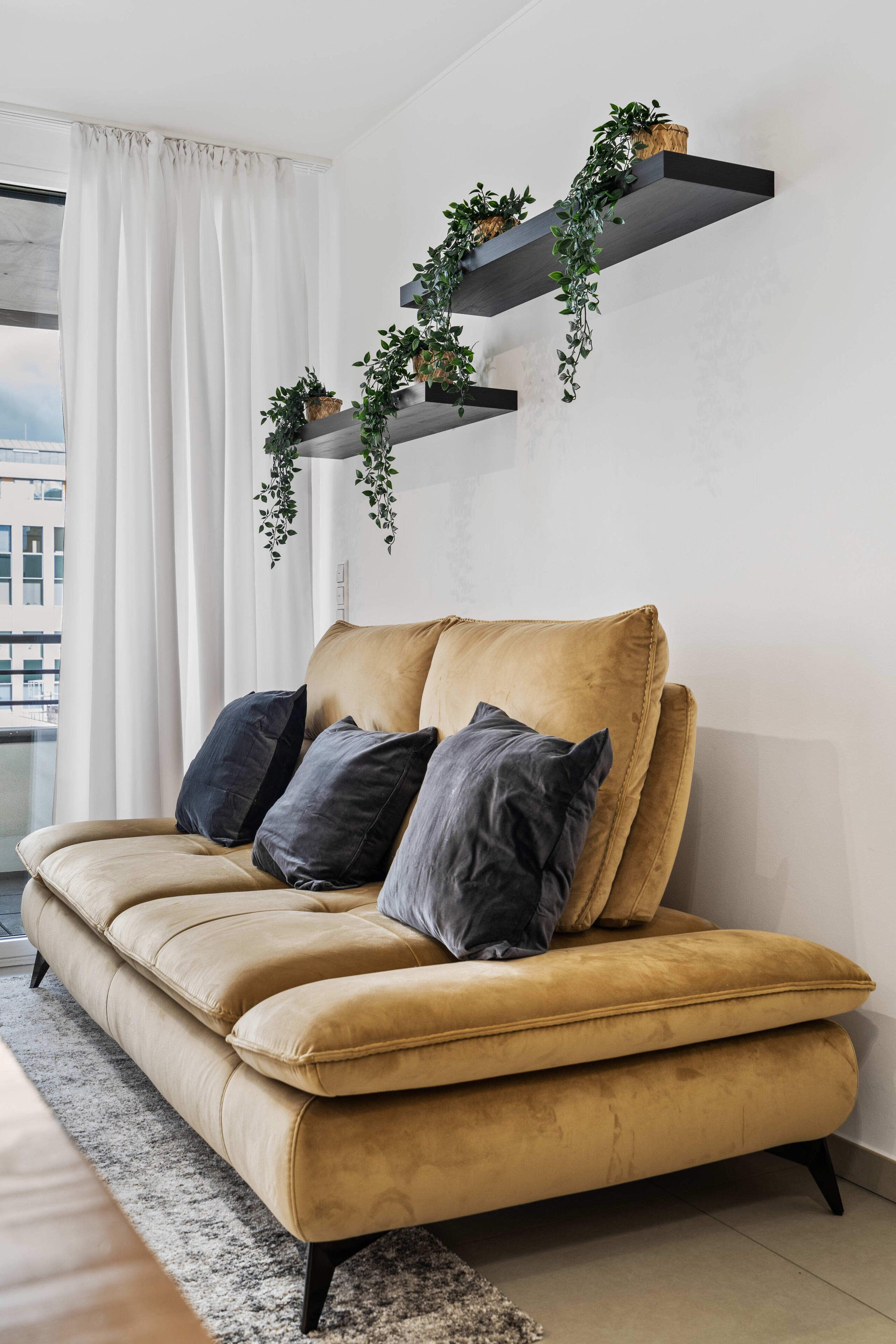 Ein senffarbenes Sofa mit dunkelgrauen Kissen, einem grauen Teppich und einem weißen Vorhang.
