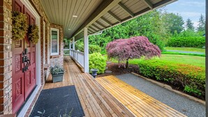 Un porche cubierto con terraza de madera, una puerta roja con detalles decorativos y un jardín cuidado con un árbol grande y arbustos.