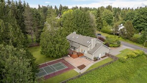 Una casa de dos pisos con cancha de tenis, rodeada de un bosque frondoso.