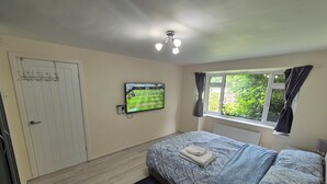Un dormitorio con cama, televisor montado en la pared, ventana con cortinas y puerta con perchero.
