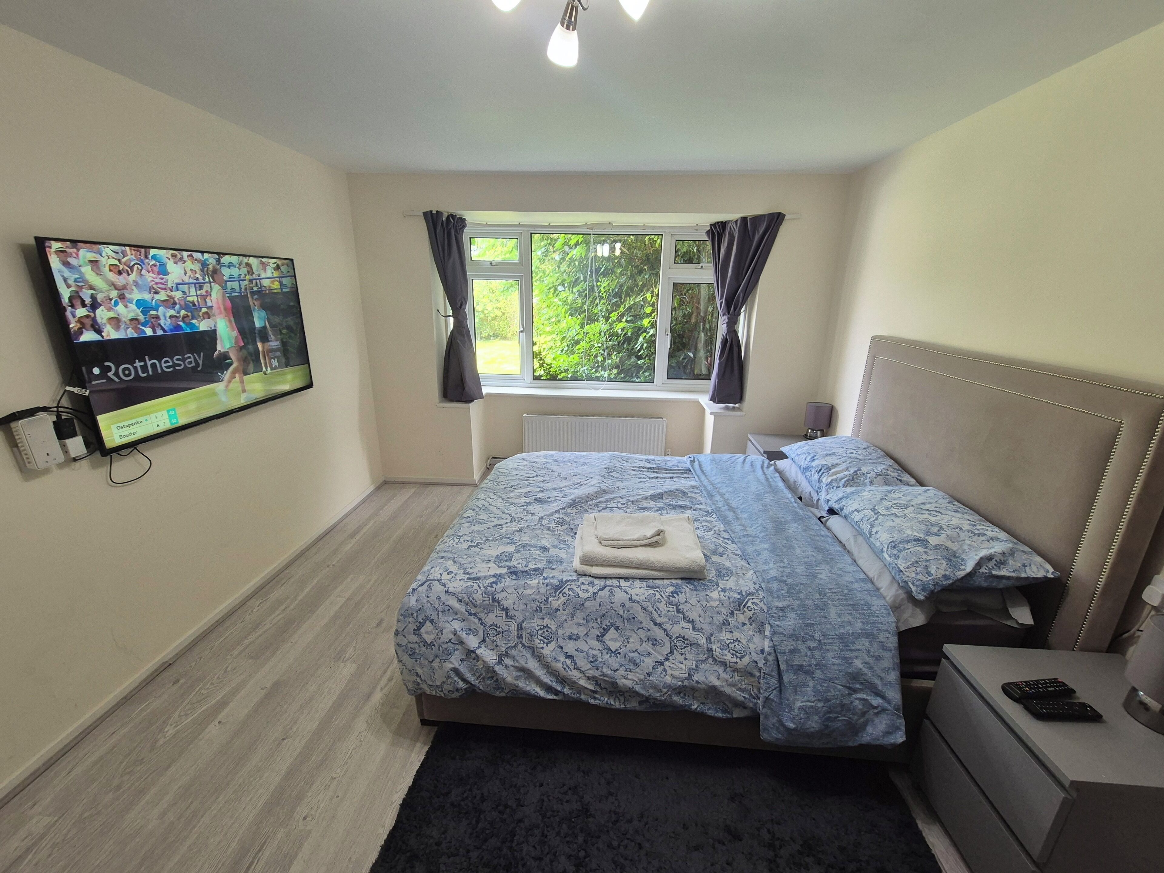 Une chambre à coucher comprenant un lit, un téléviseur fixé au mur, une fenêtre avec des rideaux, une table de chevet et une commode.