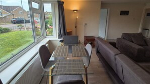 Una sala de estar moderna con una mesa de comedor de vidrio, un sofá gris y vistas a una calle residencial.