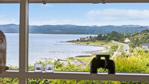 Blick vom Fenster auf die Küste, das ruhige Meer, eine Straße und einen bewaldeten Hügel.