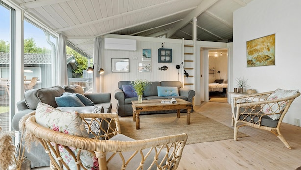 Ein geräumiges Wohnzimmer mit hoher Decke, großen Fenstern und Blick ins Freie. Es gibt Korbmöbel, ein Sofa und einen Couchtisch.