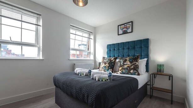 Une chambre moderne avec un grand lit, deux fenêtres, un radiateur, une tête de lit et une petite table de chevet.