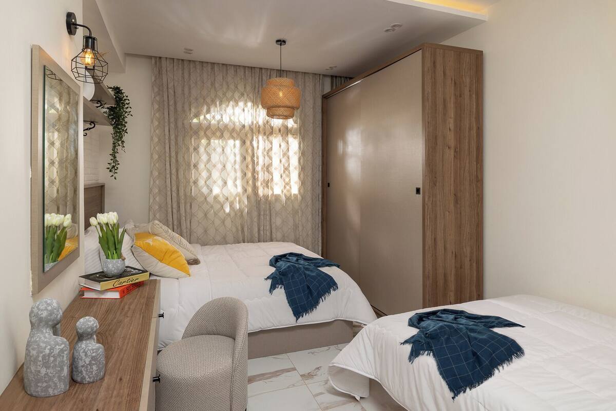 Un dormitorio moderno con dos camas, un armario de madera, un escritorio con espejo y una lámpara colgante.