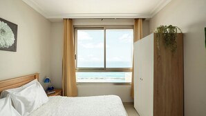 A bedroom with a large window, a bed with white linens, a wooden headboard, a nightstand with a lamp, and a view of the sea. - Carmel Seaside Sunny Stays by Sea N Rent (Haifa)