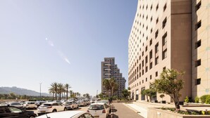 A parking lot with multiple cars, a tall building with a grid-like facade, and palm trees along the sidewalk. - Carmel Seaside Sunny Stays by Sea N Rent (Haifa)