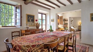 Un espace repas avec une grande table recouverte d’une nappe à motifs, des chaises en bois et un plafond rustique avec des poutres apparentes.