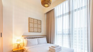 Una habitación de hotel moderna con una cama grande, una lámpara de noche, una obra de arte en la pared y un ventanal con cortinas translúcidas.