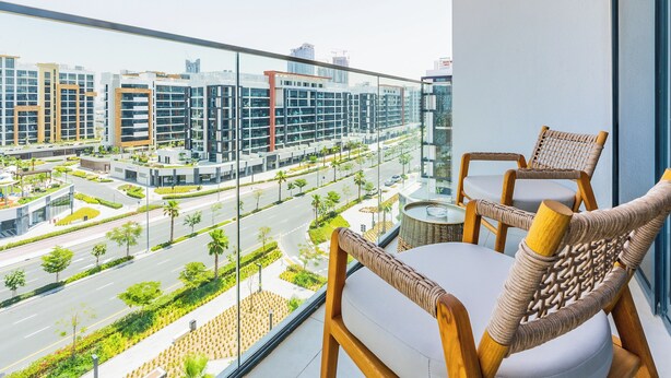 Un balcón con sillas de mimbre, barandilla de vidrio y vista a edificios modernos y una calle.