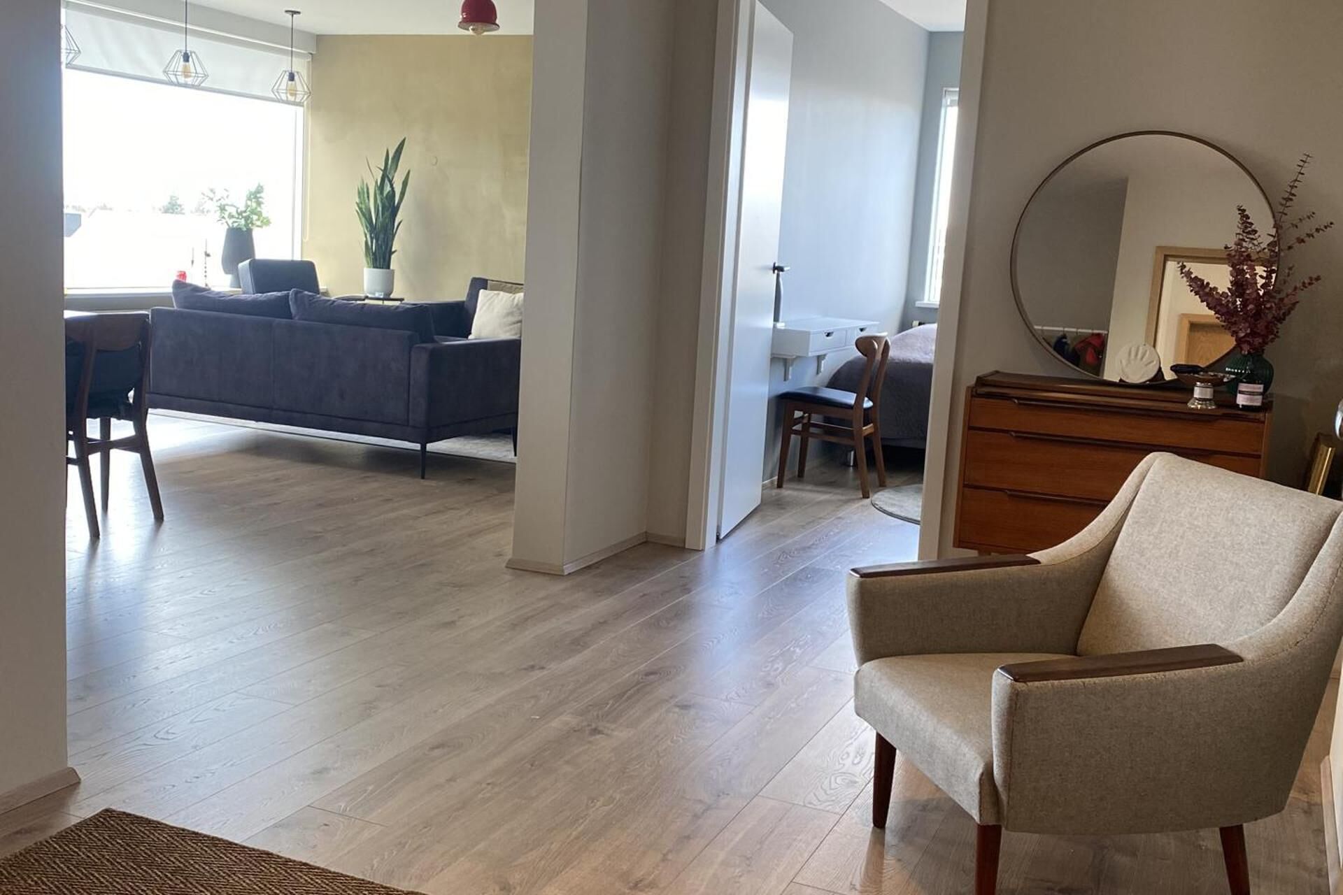 A spacious living area with a grey sofa, a wooden chair, and a small table. There is a mirror on the wall and a dresser with a vase of flowers.
