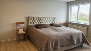 Un dormitorio con cabecera capitonada, una silla de madera, una mesita de noche y una ventana con vistas a la vegetación.