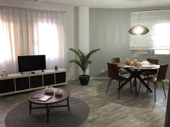 Ein modernes Wohnzimmer mit Flachbildfernseher, einem Esstisch mit Stühlen, einer Topfpflanze und einem großen Fenster mit durchsichtigen Vorhängen.
