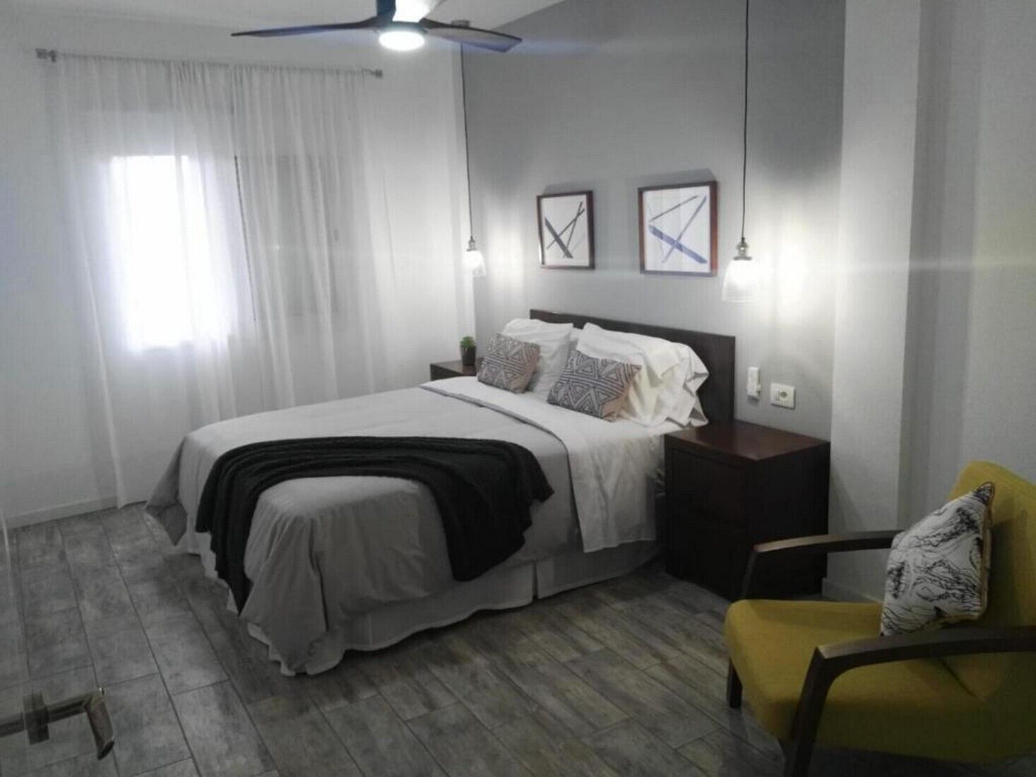 Un dormitorio con cama, mesita de noche, silla y dos luces fijadas a la pared.