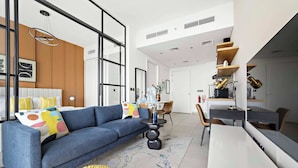 A modern living room with a blue sofa, a round yellow ottoman, and a dining area with a white table and chairs. - Silkhaus Socio Towers Dubai Hills (Dubai)