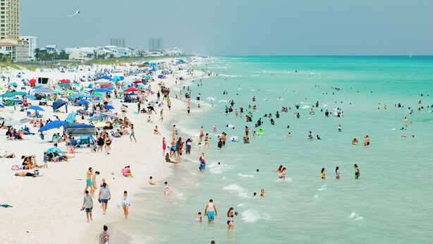 Une plage bondée où l’on voit des gens se promener et nager, ainsi que de nombreux parasols et chaises de plage.