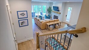 A spacious living room with a sofa, a coffee table, and a foosball table. There is a TV mounted on the wall and a staircase leading to another level. - Big Oak Bungalow by Chatuge Home Concierge | Lake House · Private Dock · Hot Tub · Ping Pong · Fireplace (Hiawasee)