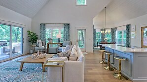A spacious living room with a high ceiling, large windows, a central seating area with a sofa and coffee table, and a kitchen with a bar area. - Big Oak Bungalow by Chatuge Home Concierge | Lake House · Private Dock · Hot Tub · Ping Pong · Fireplace (Hiawasee)