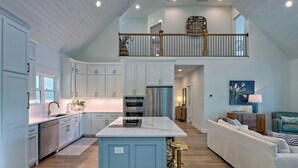 A spacious kitchen with white cabinets, a central island, and a dining area with a white sofa and a blue accent wall. - Big Oak Bungalow by Chatuge Home Concierge | Lake House · Private Dock · Hot Tub · Ping Pong · Fireplace (Hiawasee)