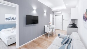 Een moderne woonkamer met een bank, een eethoek met tafel en stoelen, een flatscreen televisie aan de muur en een keuken met een koffieapparaat.