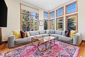 A modern living room with a gray sectional sofa, a wooden coffee table, and a colorful rug. Large windows offer views of trees. - Chateau Hadarian (Vail)