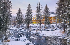 A lodge with a river in the foreground, surrounded by snow-covered trees. - Grand Hyatt|The Grand Mountain Chateau with Sweeping Views on the Cascades of Gore Creek (Vail)
