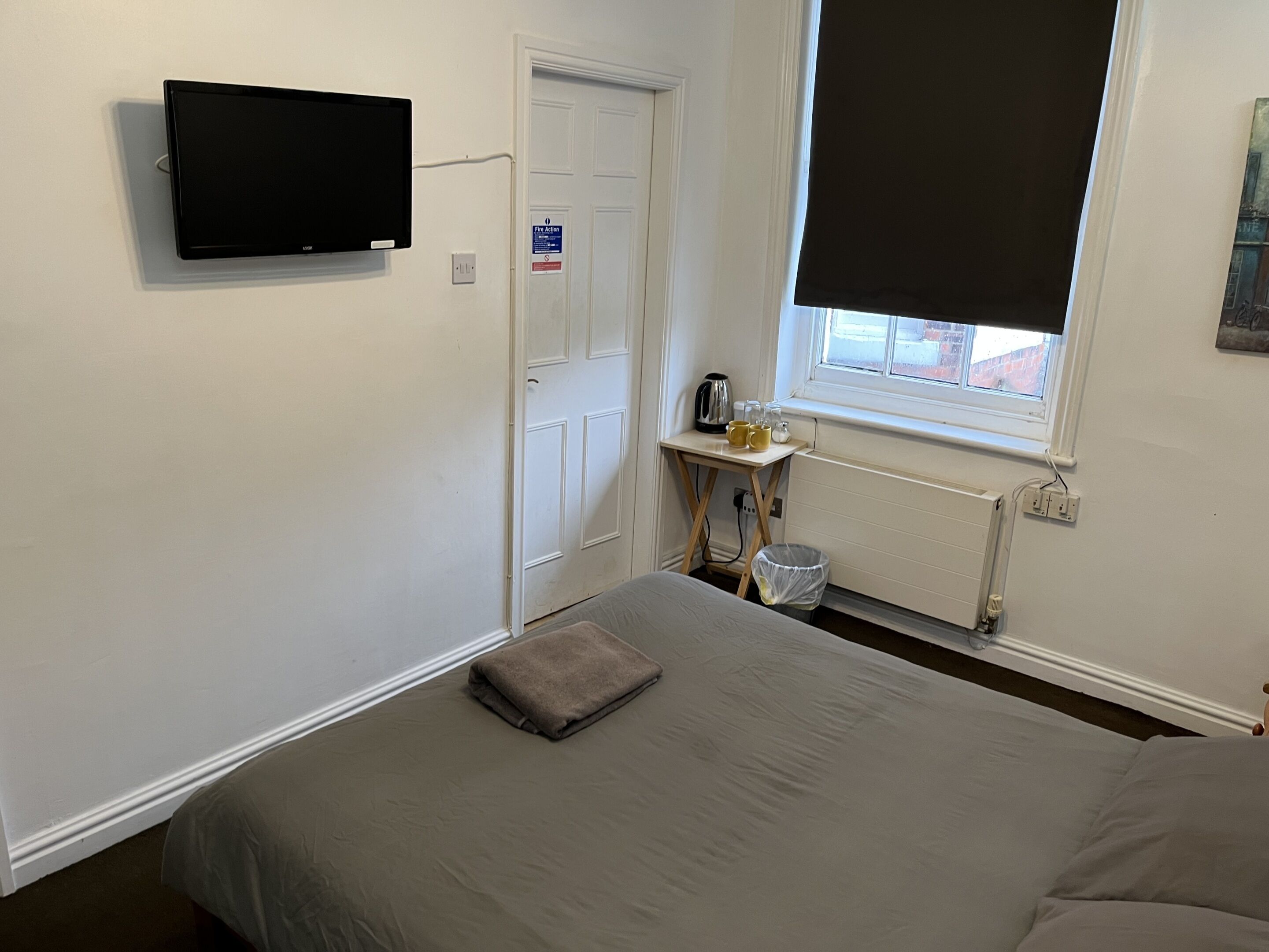 A bedroom with a bed, a TV mounted on the wall, a small table with a kettle and cups, a window with a blind, and a door with a fire action sign.