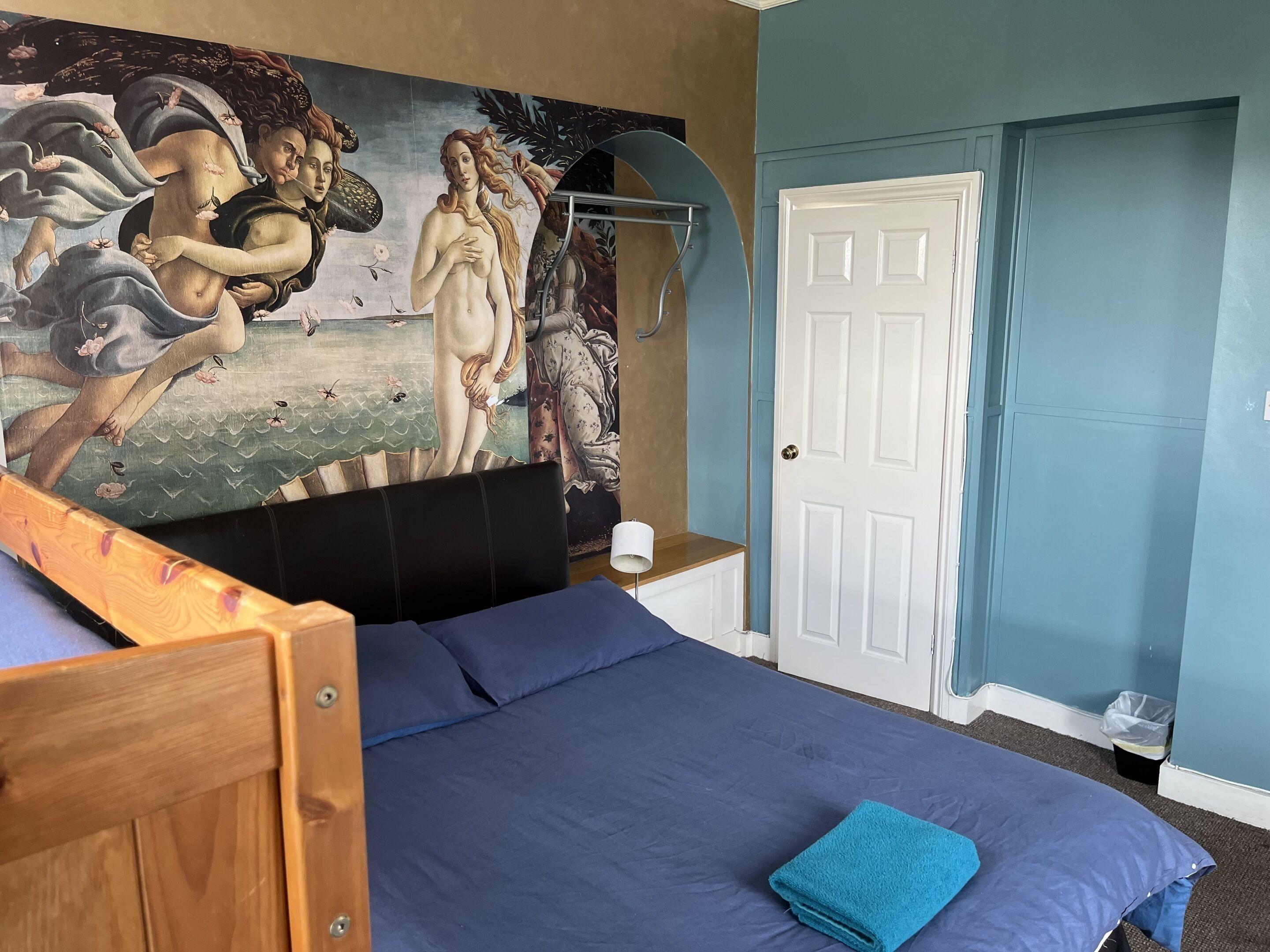 A bedroom with a large mural, a bed with a blue bedspread, a wooden headboard, and a white door.