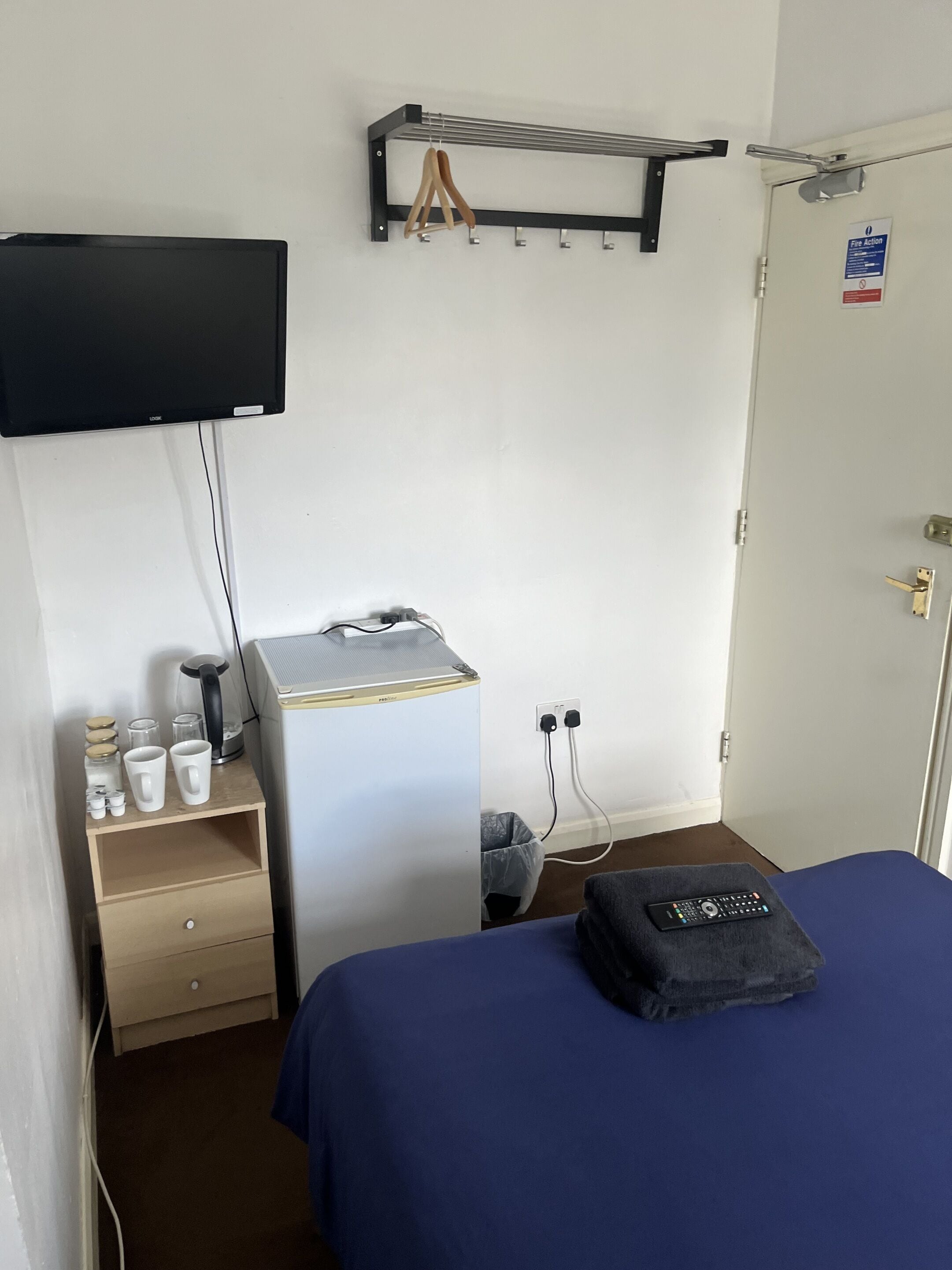 Habitación de hotel con cama, escritorio con cafetera, televisor montado en la pared y puerta con señal de procedimiento de emergencia.