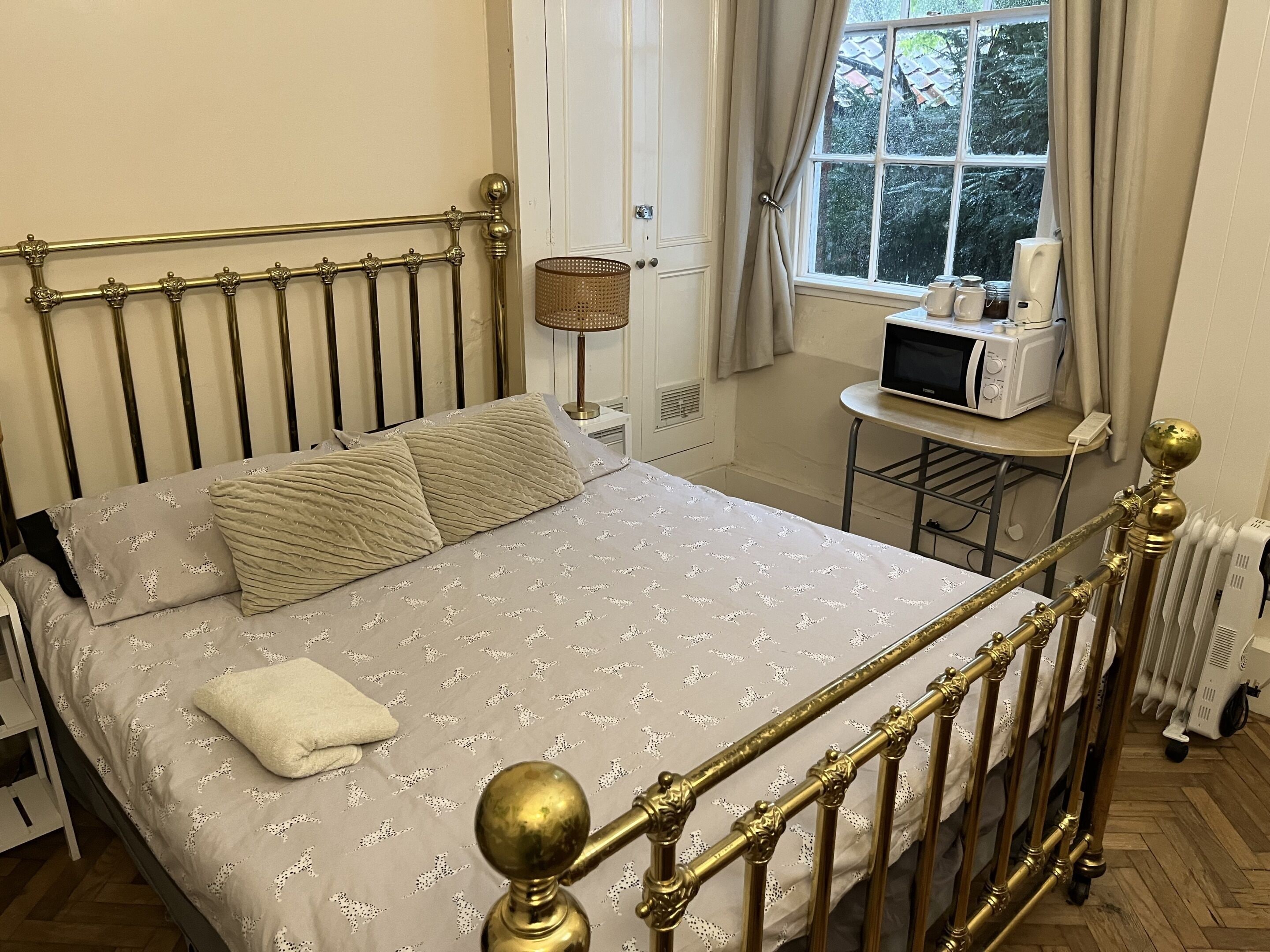 A neatly made bed with a patterned bedspread and pillows, a bedside table with a lamp and a microwave, and a window with curtains.