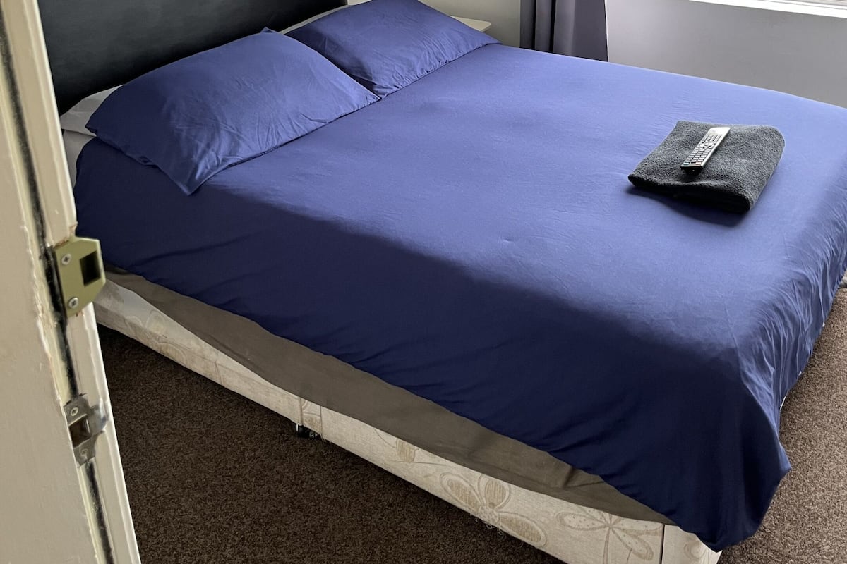 A single bed with a black headboard and a blue bedspread, a remote control on the bed, and a window with a view of a grassy area.