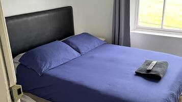 A single bed with a black headboard and a blue bedspread, a remote control on the bed, and a window with a view of a grassy area.