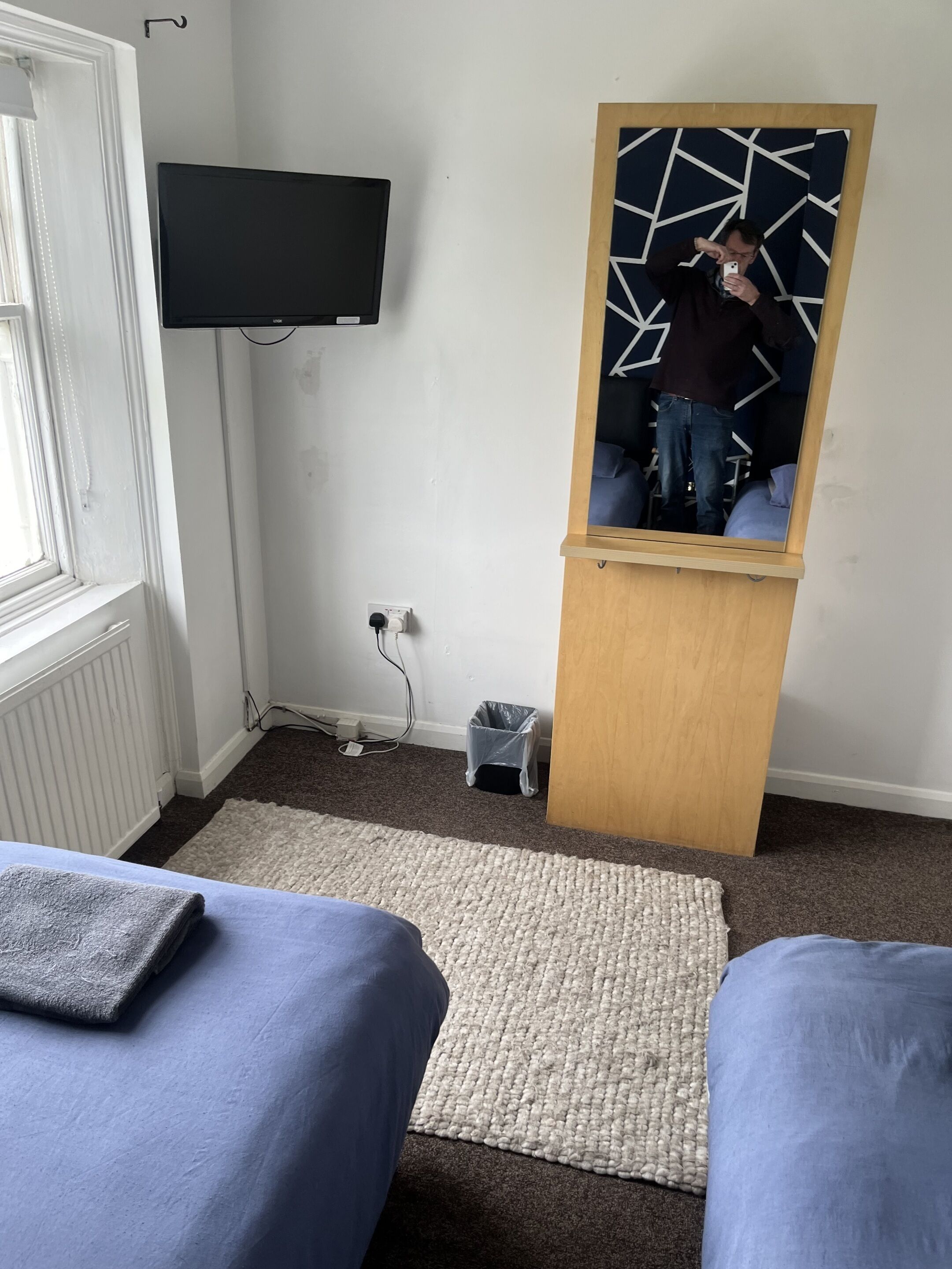 Un dormitorio con dos camas, un televisor montado en la pared, un espejo con diseño geométrico y un pequeño cubo de basura.