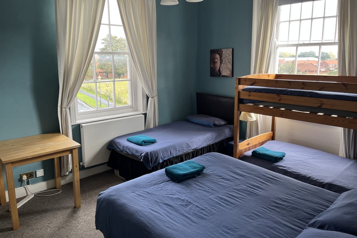 A dormitory room with two beds, a wooden table, and a window with curtains.