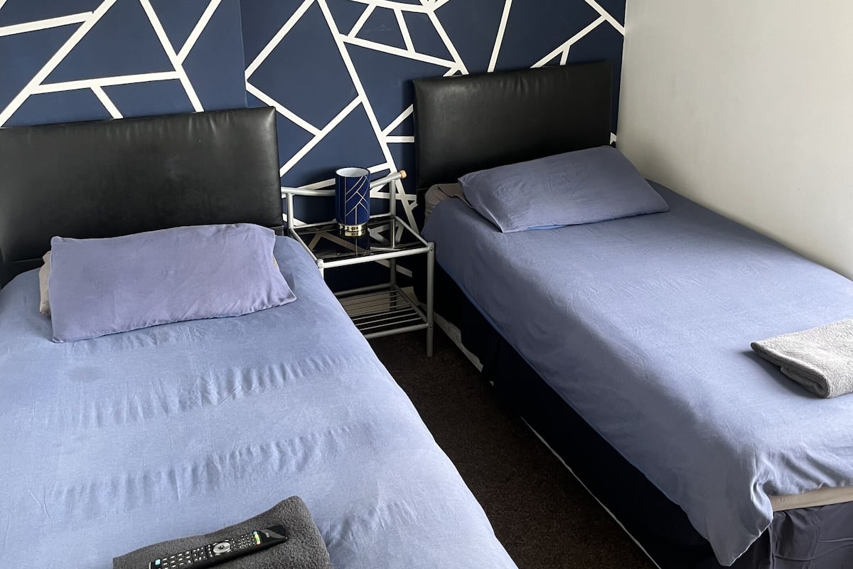 A hotel room with two beds, a geometric-patterned wall, a small table with a cup, and a remote control on one bed.