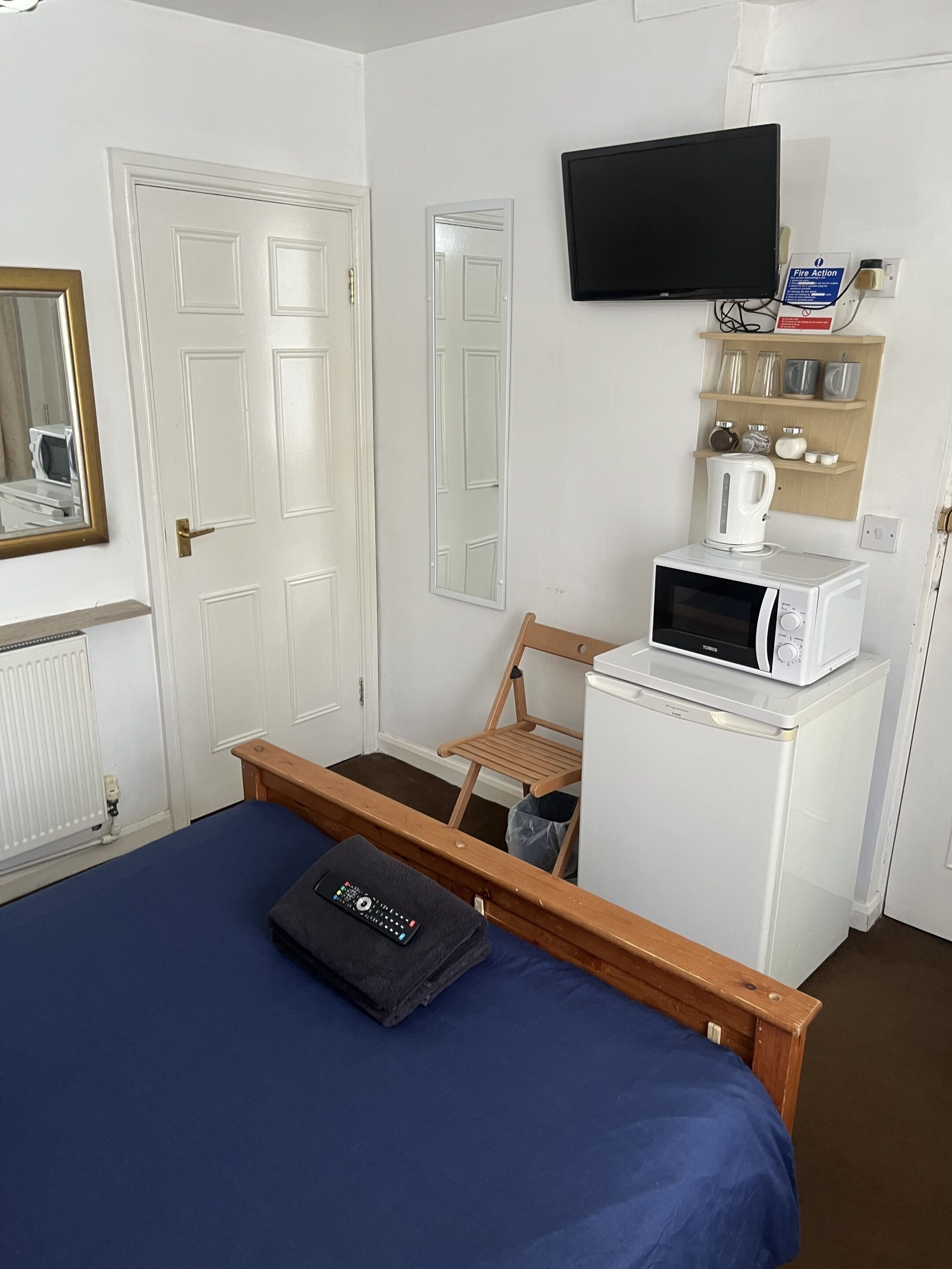 A compact hotel room with a bed, a wooden chair, a microwave, a television, and a shelf with mugs and a kettle.
