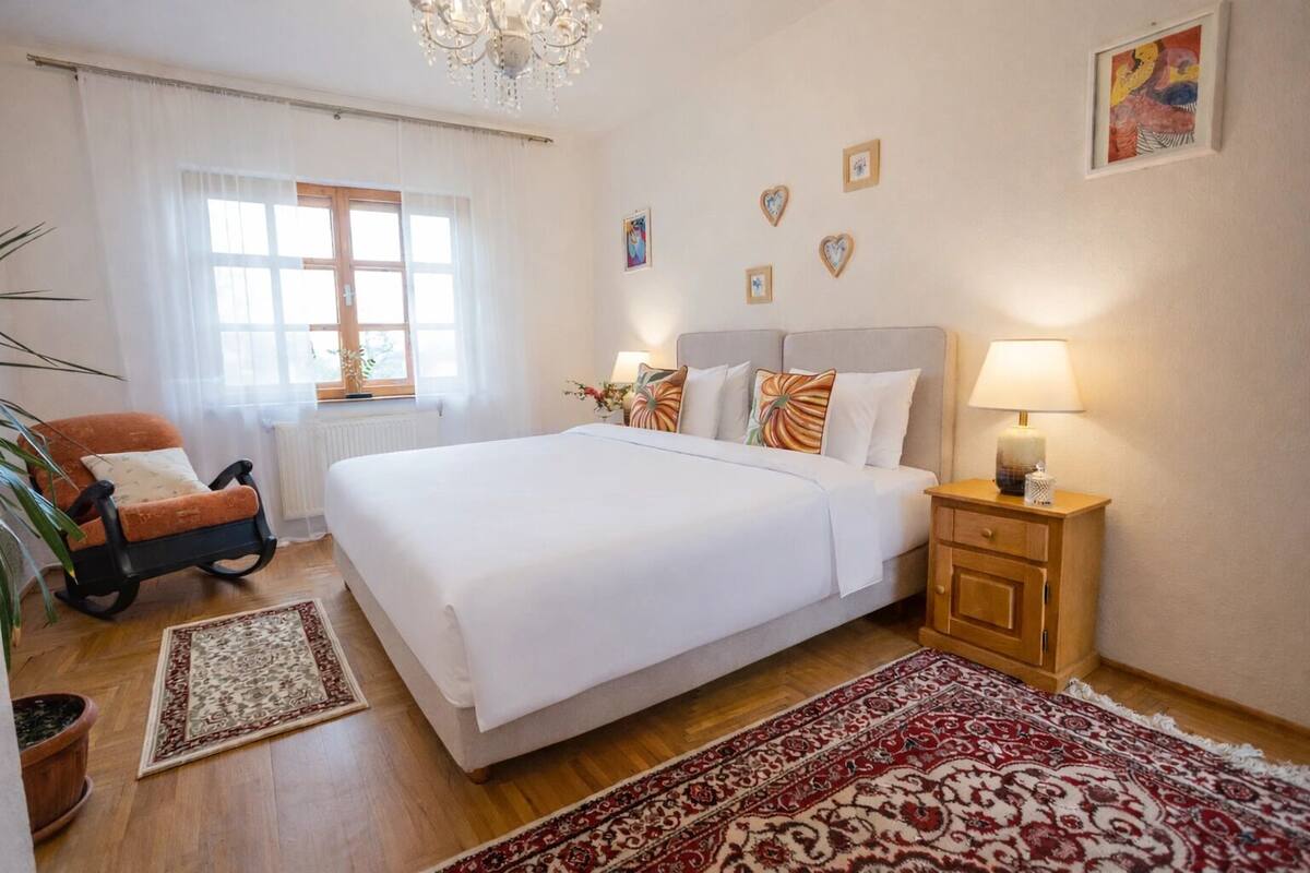 Un dormitorio con una cama grande, una mesita de noche, una silla, una ventana con cortinas y un candelabro.
