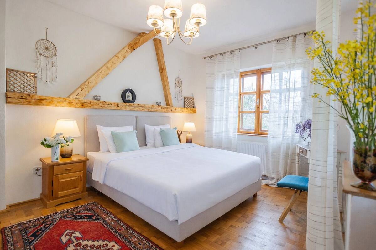 Un dormitorio con cama, cabecera de madera, mesitas de noche, una mesilla auxiliar, un tapete rojo estampado, una ventana con cortinas y un florero con flores.
