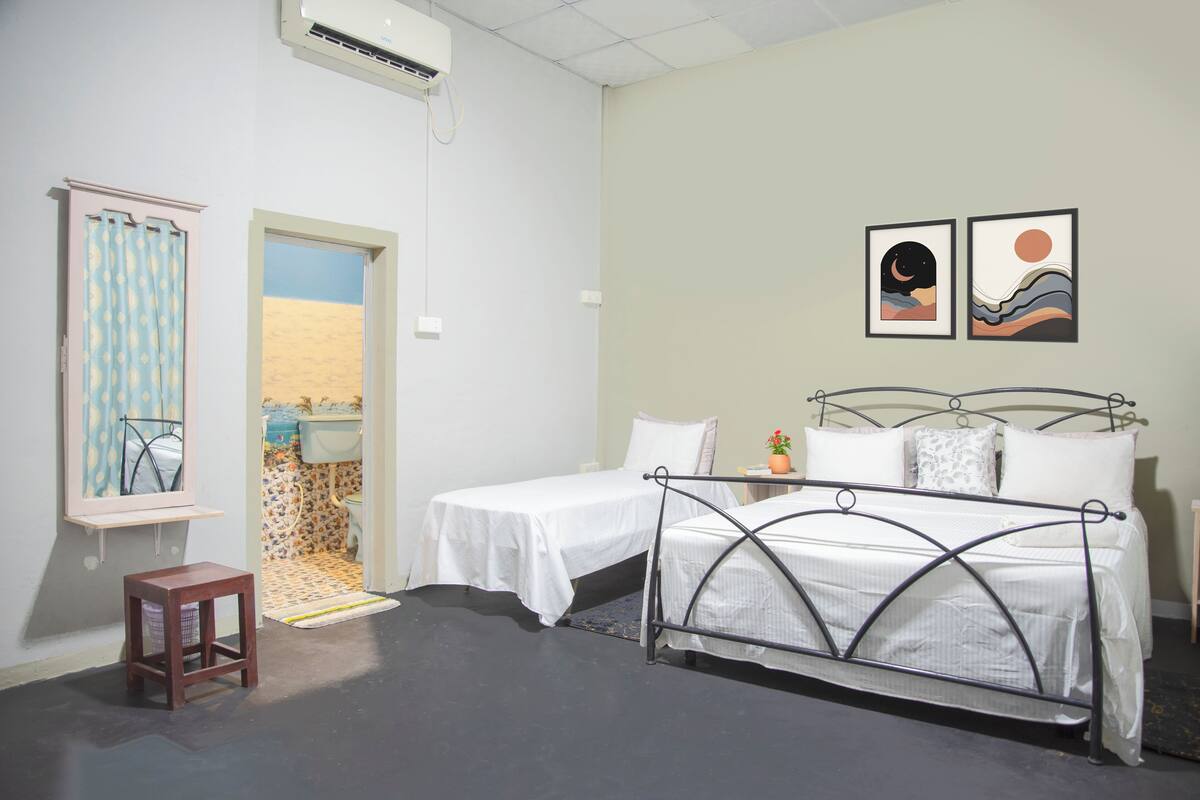 A room with two single beds, a small wooden stool, a window with a curtain, a sink, and two framed artworks on the wall.