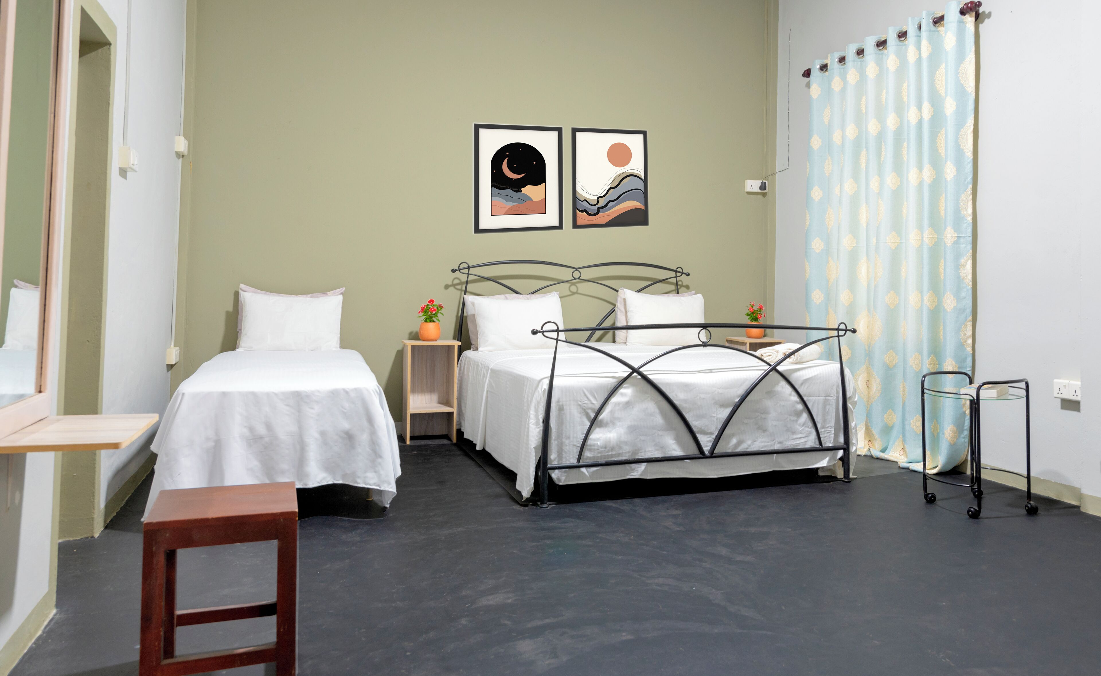 Un dormitorio con dos camas individuales, una mesita, una mesilla de noche, estructura de cama metálica, una planta en maceta y una cortina estampada.