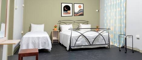 Un dormitorio con dos camas individuales, una mesita, una mesilla de noche, estructura de cama metálica, una planta en maceta y una cortina estampada.