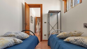 A narrow hallway with two beds, each with matching pillows and blue bedding. A metal rack is mounted on the wall, and a door leads to another room.