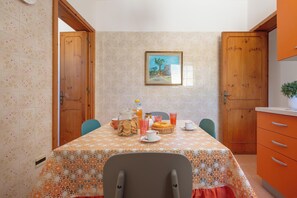 A dining area with a table set for two, featuring a floral tablecloth, a jar of cookies, a teapot, and a fruit bowl.