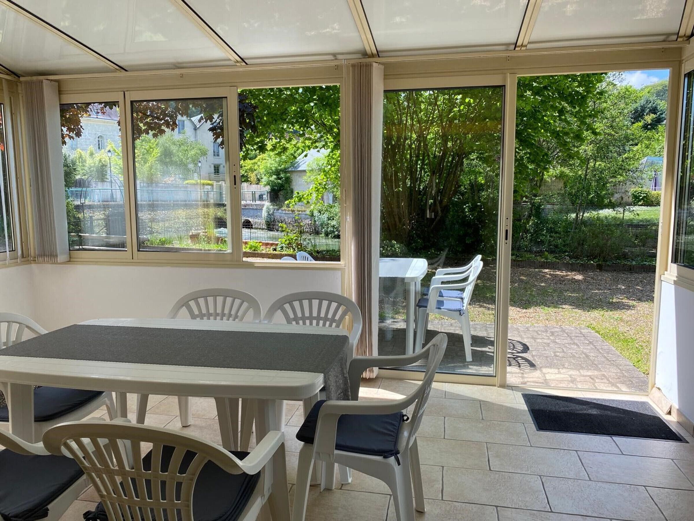 A covered patio area with a table and chairs, large windows offering views of a garden and a building.