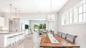 A spacious kitchen with a long wooden dining table, white bar stools, pendant lights, and a view of the outdoors.