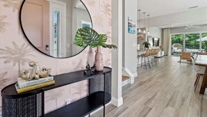 A modern living room with a large round mirror, a console table with decorative items, a dining area with wooden chairs, and a view of the outdoors.